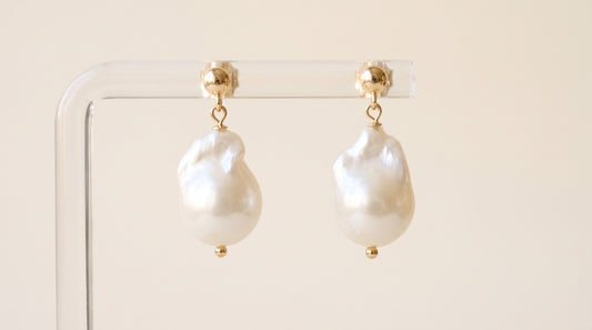 A pair of baroque pearl drop earrings with gold posts, hanging from a clear acrylic display stand against a warm beige background.