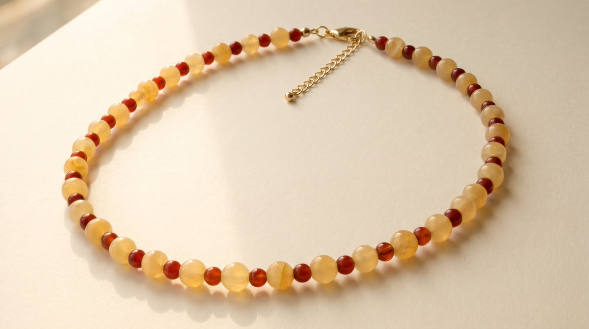 A beaded necklace made of translucent pale yellow and dark red round beads, with a gold-tone chain extender and clasp, displayed on a light cream surface.