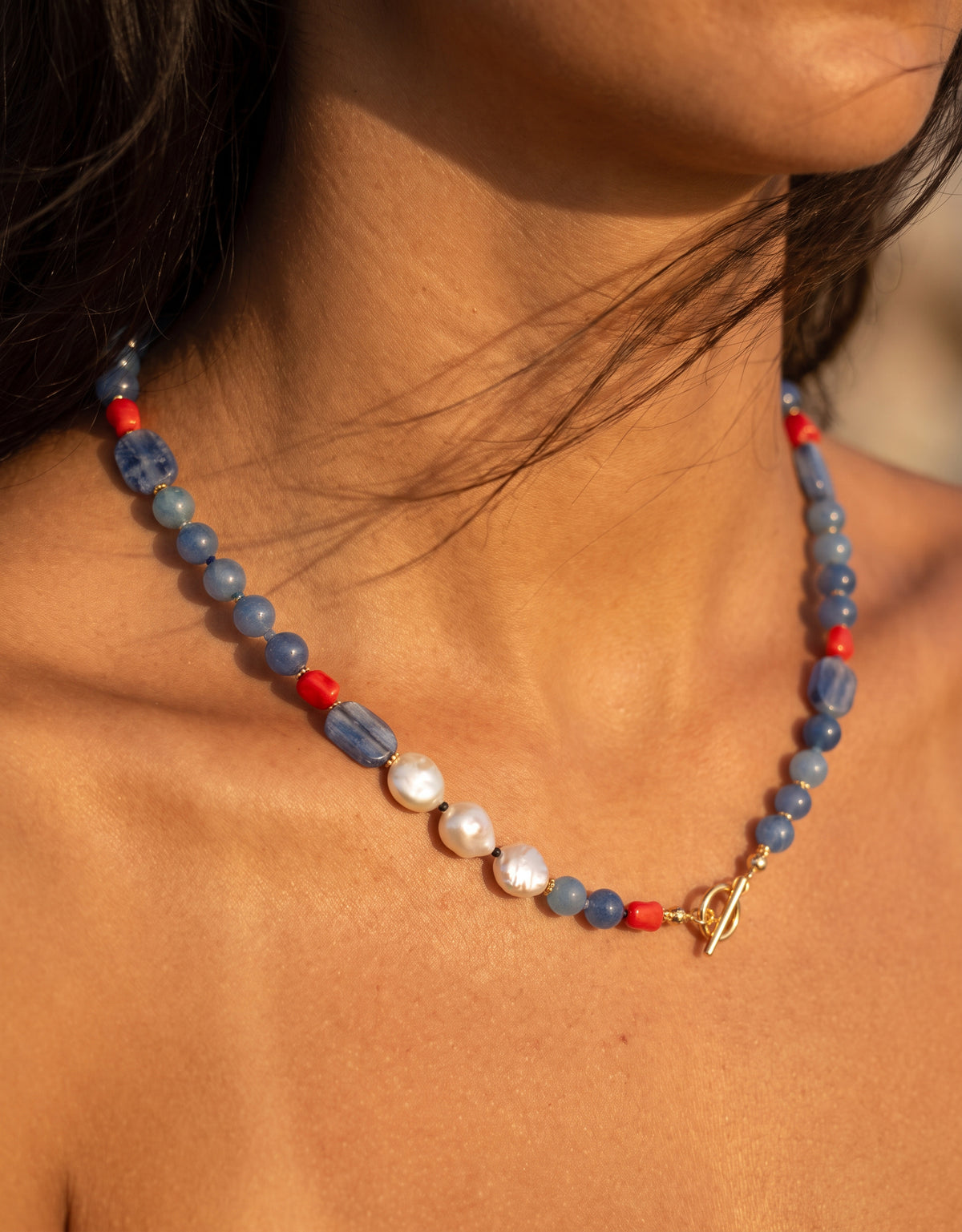 A close-up of a woman wearing a beaded necklace with blue stone beads, red accent beads, white pearls, and a gold toggle clasp.