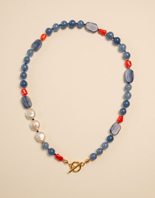 A beaded necklace featuring light blue stone beads, polished red cylindrical beads, irregular white faux pearls, and elongated blue-gray stone accents, finished with a gold-tone toggle clasp, displayed on a warm beige background.