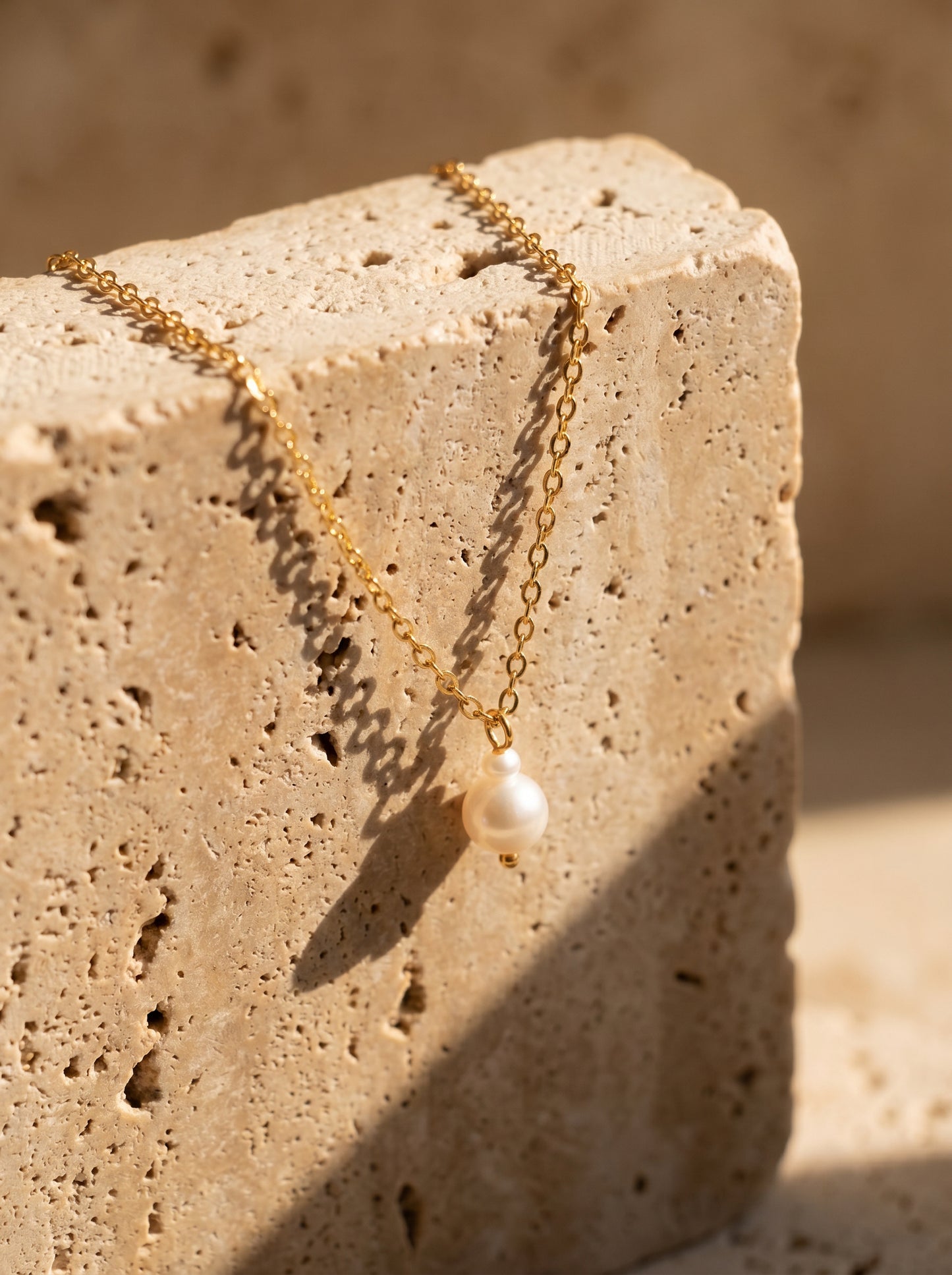 A delicate gold chain necklace with a single white freshwater pearl pendant, draped over a textured beige travertine stone block in warm, direct sunlight.