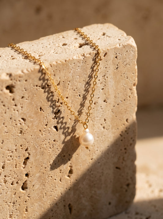 A delicate gold chain necklace with a single white freshwater pearl pendant, draped over a textured beige travertine stone block in warm, direct sunlight.