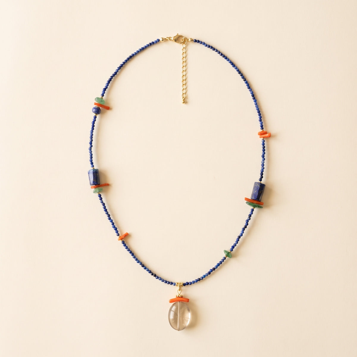 A beaded necklace with a clear teardrop pendant, featuring dark blue beads, orange and green accent beads, and gold-toned metal hardware, displayed on a light beige background.