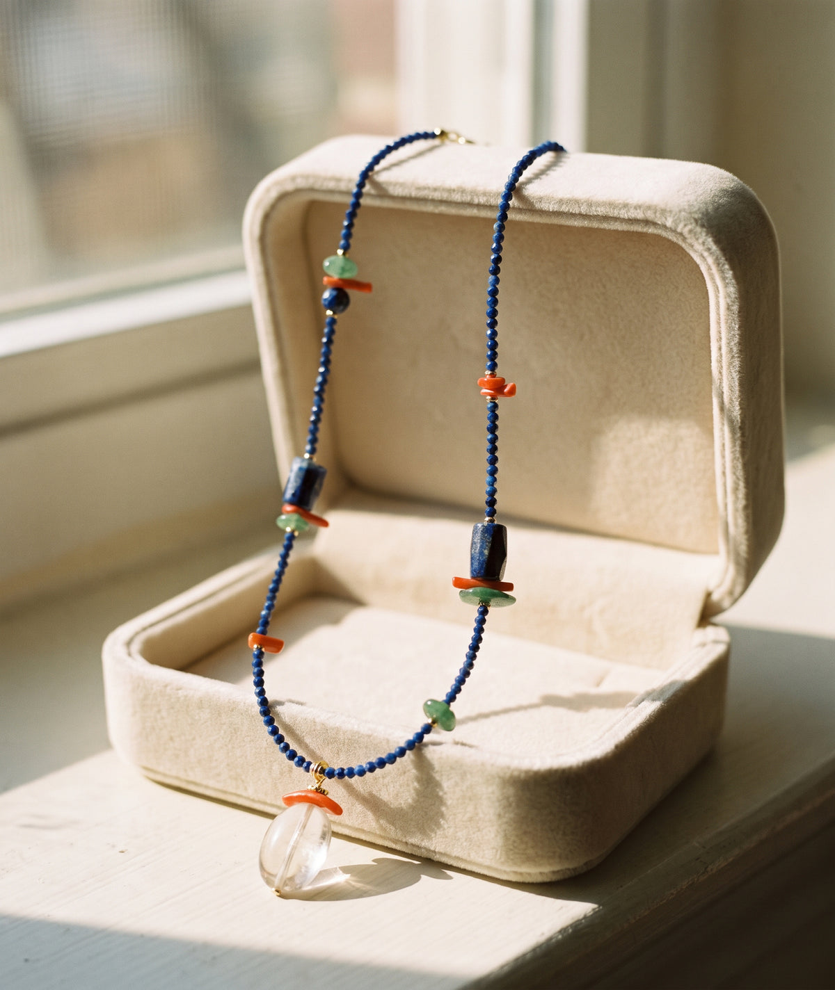 A beaded necklace with dark blue beads, orange coral-like accents, light green beads, and a clear faceted pendant, displayed inside an open cream-colored jewelry box on a windowsill.