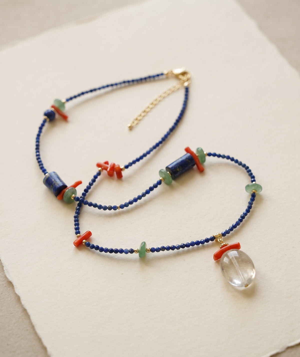 A delicate beaded necklace with dark blue beads, coral-red spacer accents, light green beads, and a clear faceted oval pendant, displayed on a textured cream paper background.