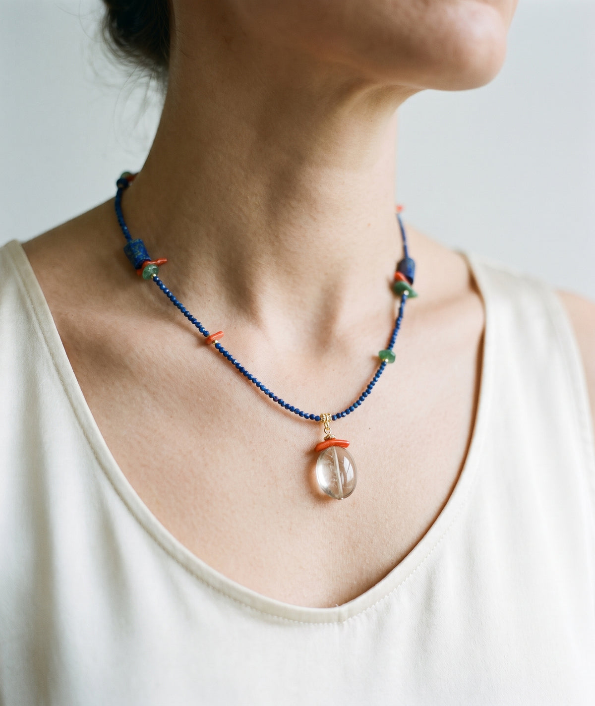 A close-up of a person wearing a beaded necklace with a clear teardrop-shaped pendant. The necklace features dark blue beads with small orange and green accents.