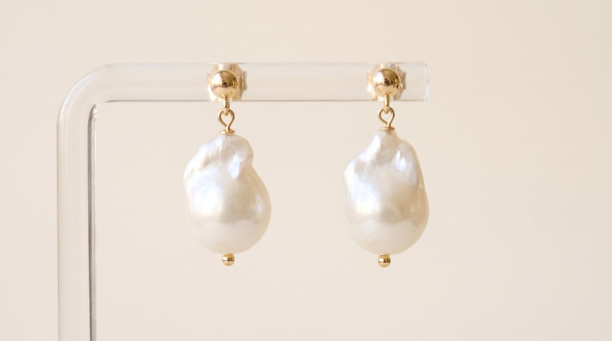 A pair of baroque pearl drop earrings with gold posts, hanging from a clear acrylic display stand against a warm beige background.