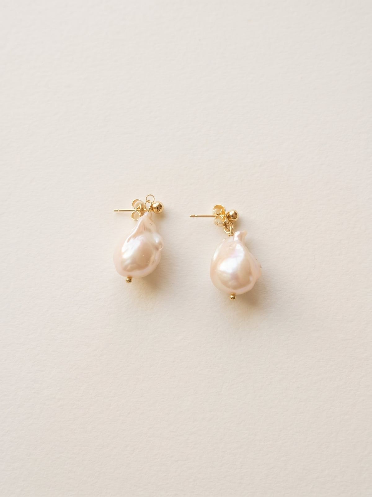 A pair of gold stud earrings featuring irregular, teardrop-shaped pale pink baroque pearls, displayed on a clean, light beige textured background.