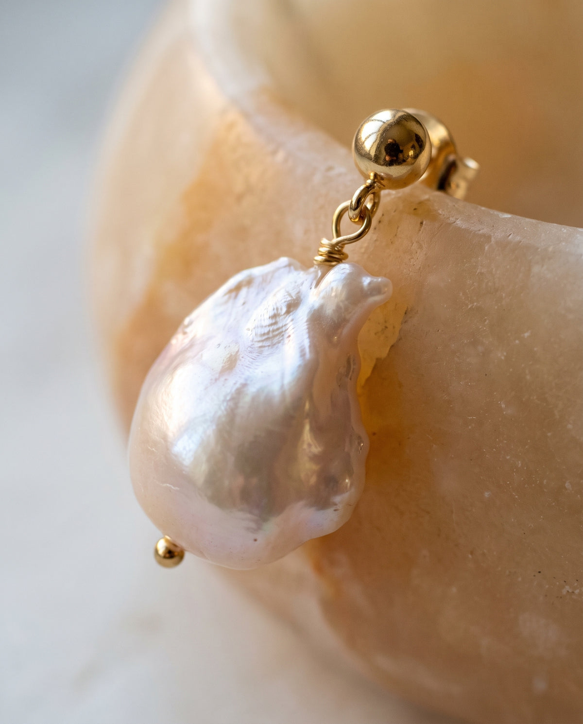 A close-up of a gold hoop earring with a large, irregular white baroque pearl dangling from it, resting against a smooth, warm beige stone surface.