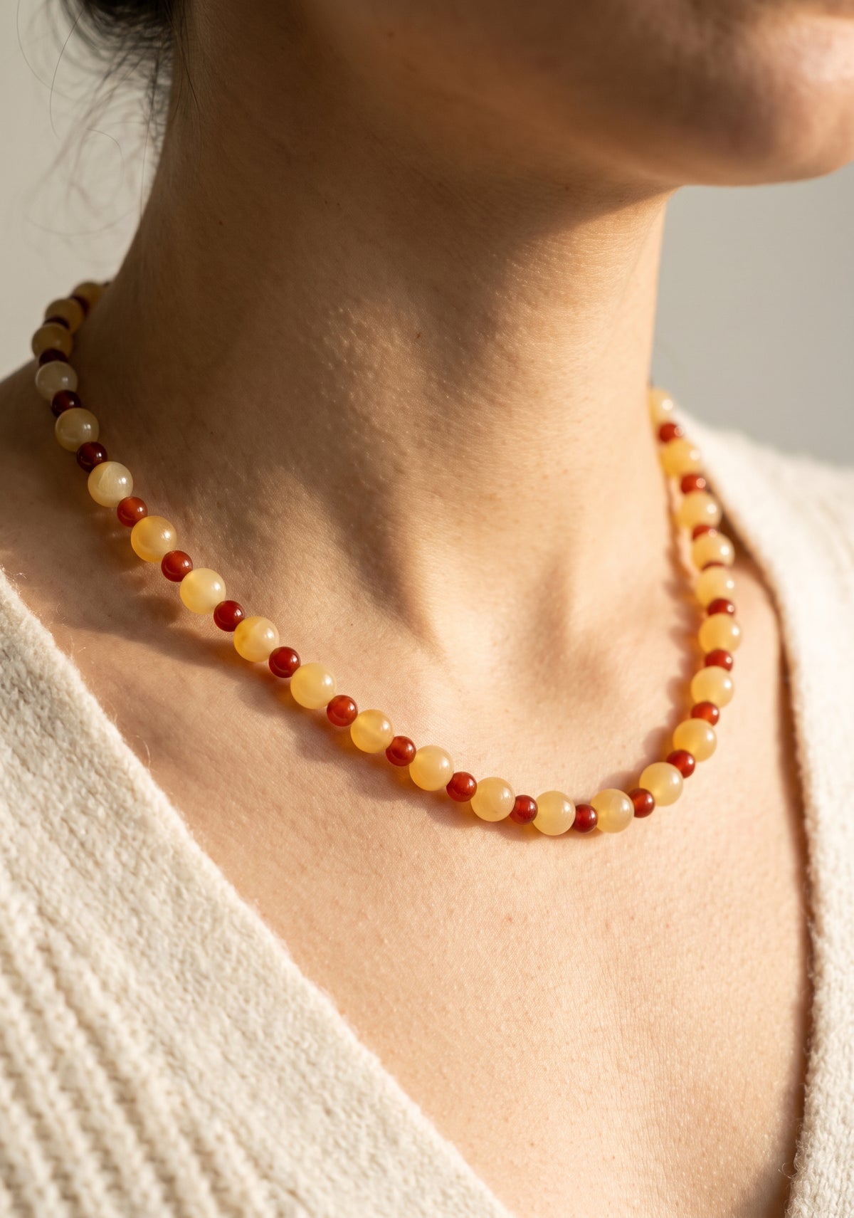 A close-up of a person wearing a beaded necklace made of alternating translucent pale yellow and deep red round beads, paired with a cream knit top.