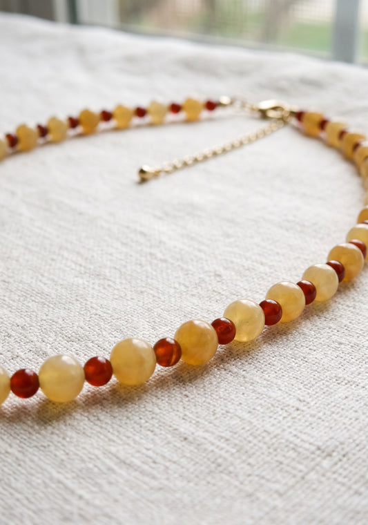 A beaded necklace with alternating translucent pale yellow and deep red round beads, accented by a gold-tone chain extender and clasp, displayed on a textured off-white linen surface.