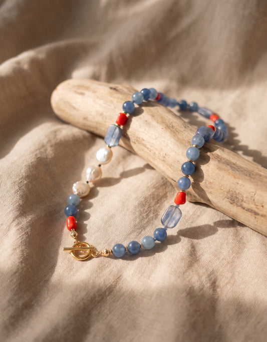 A beaded necklace with light blue and white beads, small red accents, and gold-toned metal details, draped over a piece of driftwood on a beige fabric background.