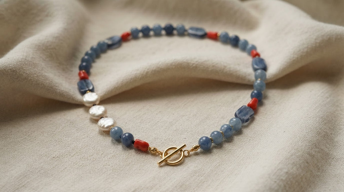 A beaded bracelet featuring blue stone beads, small red beads, white baroque pearls, and a gold toggle clasp, displayed on a draped beige fabric background.