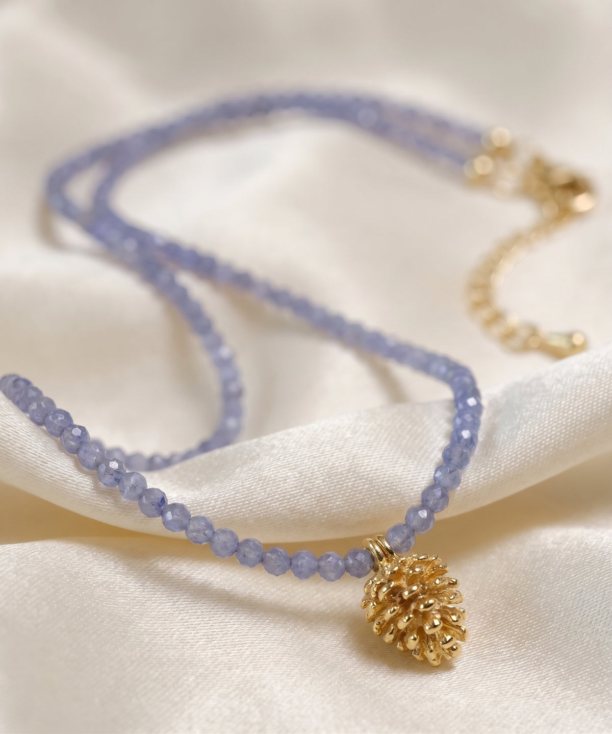A delicate double-strand necklace with light periwinkle blue faceted beads and a textured gold pinecone pendant, displayed on draped cream-colored satin fabric.