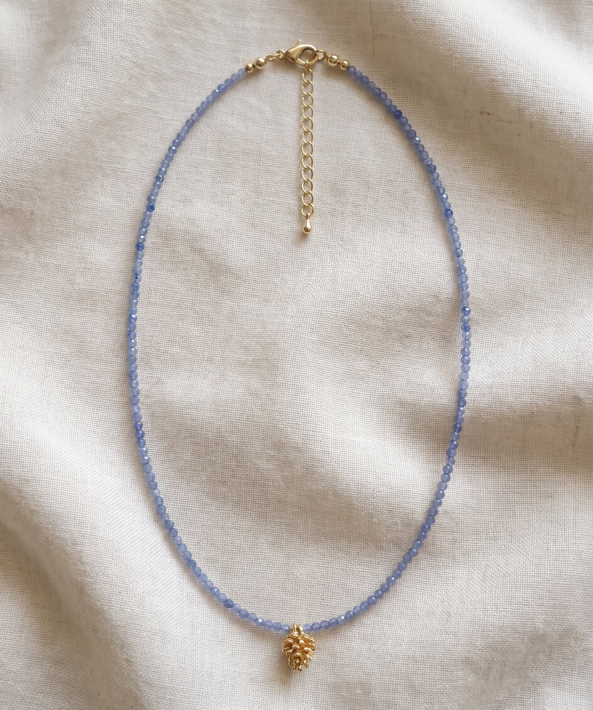A delicate beaded necklace with light blue beads and a small gold pinecone charm, displayed on a textured beige fabric background.