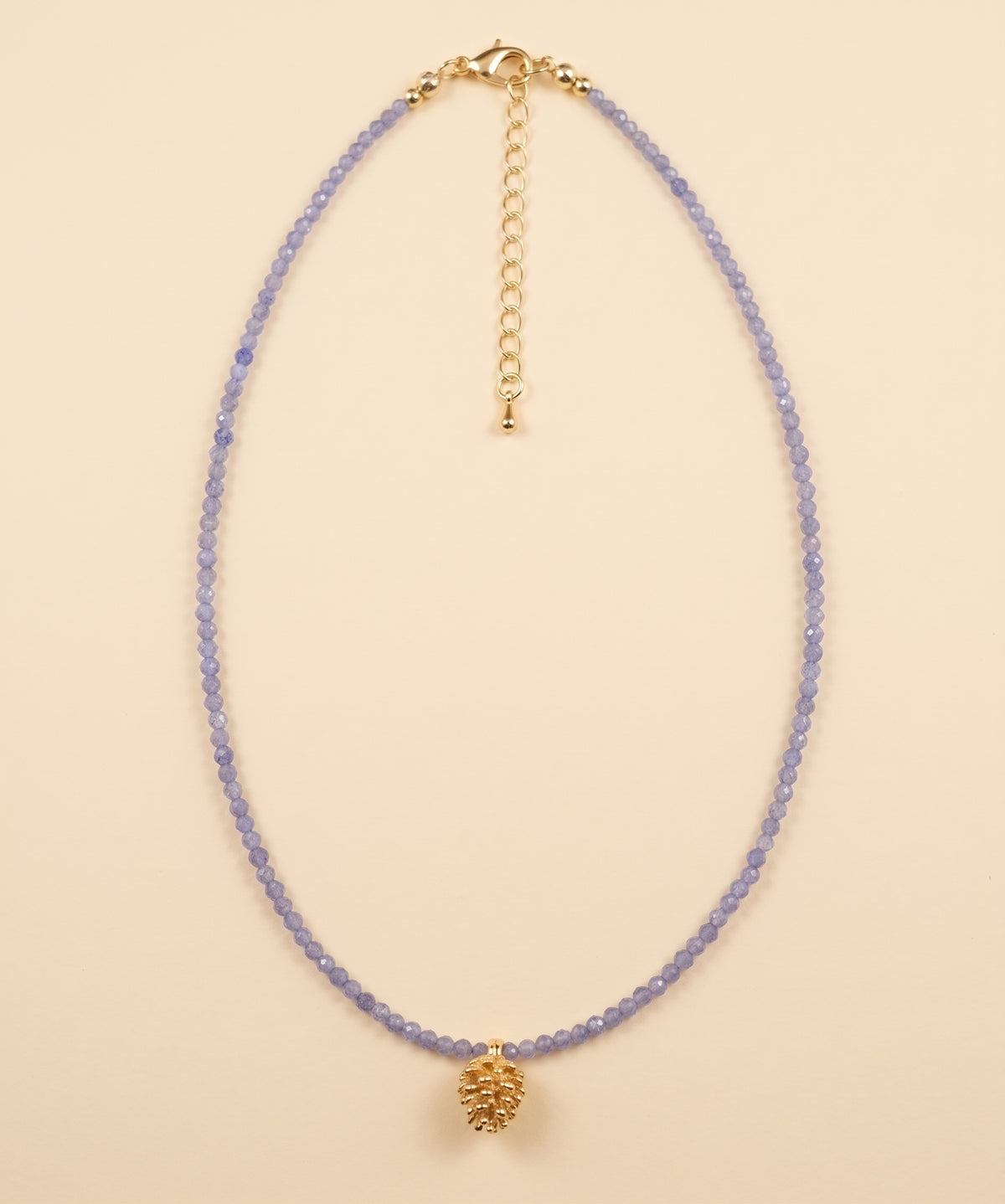A delicate beaded necklace featuring light purple beads and a textured gold pinecone-shaped pendant, displayed on a solid beige background.